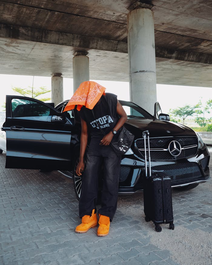 Person with a suitcase under a bridge, beside a black luxury car showcasing travel.
